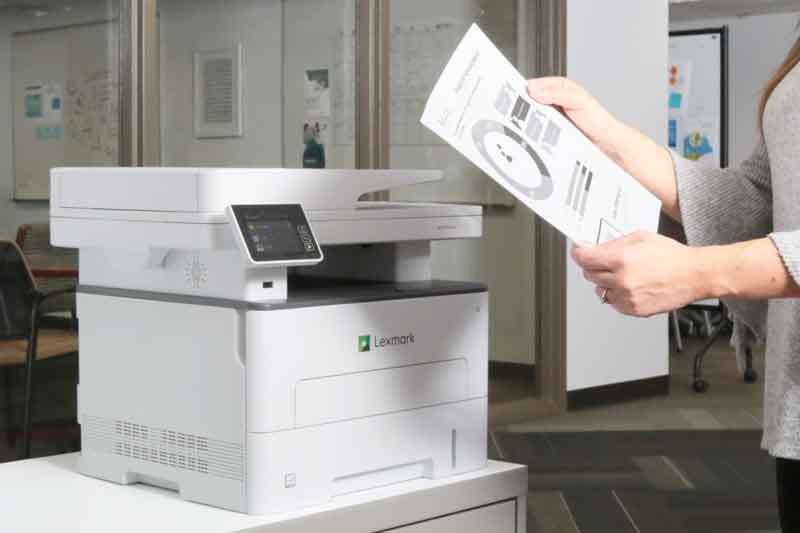 Person examines recently printed document near printer. 